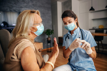 Senior woman with face mask on sitting at home and rejecting to get vaccine for corona virus. Nurse explaining her why it's better to get it.