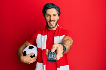 Young hispanic man football hooligan holding ball using tv control sticking tongue out happy with...