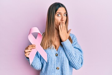 Brunette young woman holding pink cancer ribbon covering mouth with hand, shocked and afraid for mistake. surprised expression