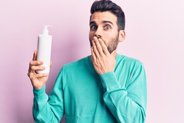Young handsome man with beard holding bottle of cream covering mouth with hand, shocked and afraid...