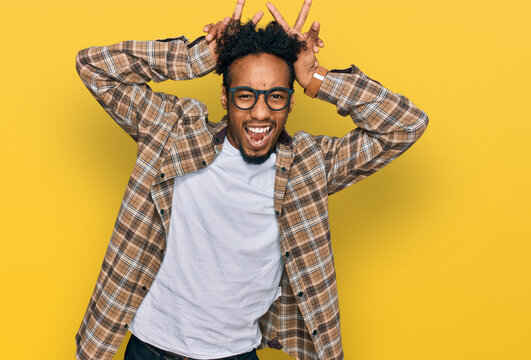 Young african american man with beard wearing casual clothes and glasses posing funny and crazy with fingers on head as bunny ears, smiling cheerful