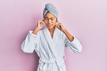 Young chinese woman wearing shower towel cap and bathrobe covering ears with fingers with annoyed expression for the noise of loud music. deaf concept. - obrazy, fototapety, plakaty
