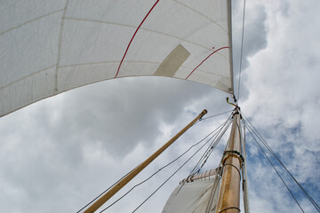 Fototapeta premium Looking up the mast of a gaff rigged sailing yacht: white sails, wooden mast and gaff, rope shrouds, halyards and sheets, and blue cloudy sunny sky