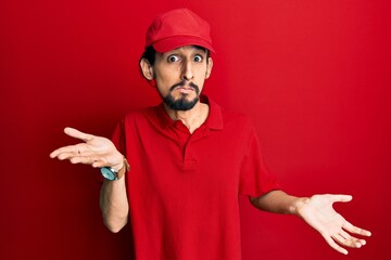 Young hispanic man wearing delivery uniform and cap clueless and confused expression with arms and hands raised. doubt concept.