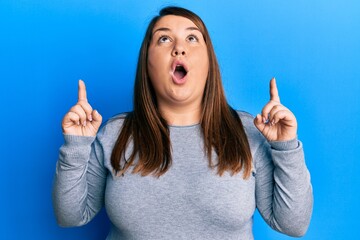 Beautiful brunette plus size woman wearing casual clothes amazed and surprised looking up and pointing with fingers and raised arms.