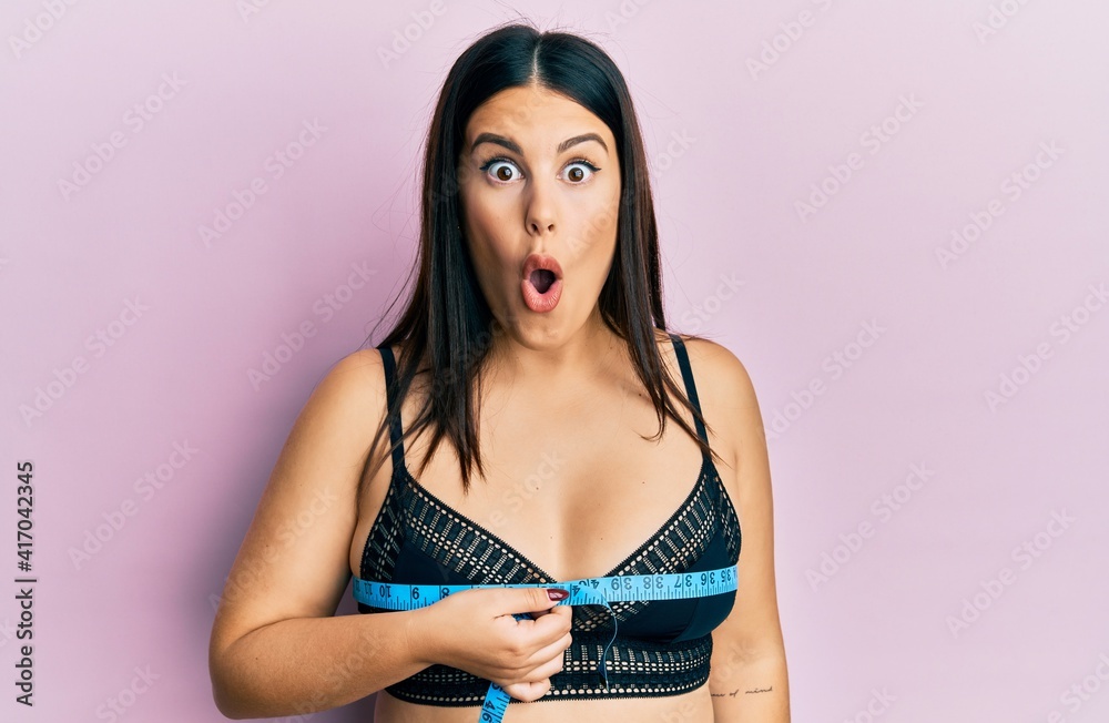 Wall mural Beautiful brunette woman using tape measure measuring breast scared and amazed with open mouth for surprise, disbelief face