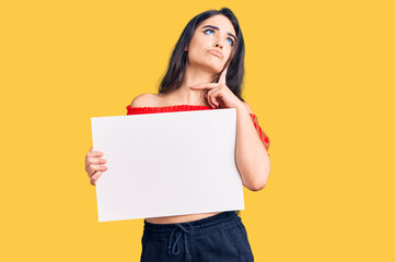 Fototapeta na wymiar Brunette teenager girl holding blank empty banner serious face thinking about question with hand on chin, thoughtful about confusing idea