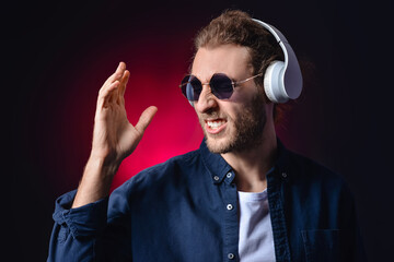 Obraz premium Young man in sunglasses and headphones enjoying music with energetic expression and raised hand on dark background, set against a dark background with a red gradient