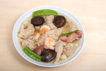 Close up view from above of a white porcelain bowl full of Wonton Soup on light brown table.