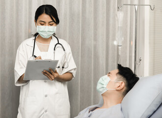 Male Asian patient wearing protective hygiene mask laying on bed and talk to female doctor that holding medical chart and writing the information of taking patient history