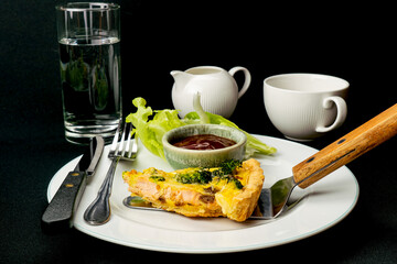 Putting a portion of freshly baked broccoli salmon quiche in white ceramic dish on black background with green vegetable and a bowl of ketchup.