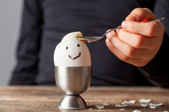 A Small Child Is Eating Soft Boiled Humpty Dumpty Egg Placed In Egg Cup On A Wooden Breakfast Table. She Uses Spoon To Eat The Egg From Its Shell. She Has Drawn A Smiling Face On The Shell.