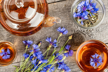 Fototapeta na wymiar Cups of herbal tea, transparent teapot and blue cornflowers flowers on wood background. Top view Flat lay.