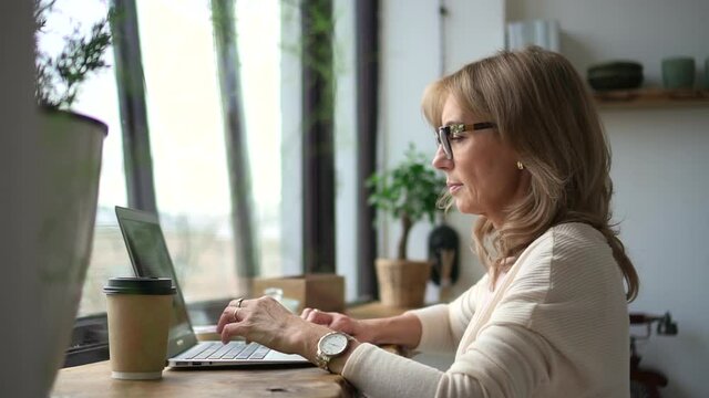 Businesswoman working with laptop at table and staying at home during pandemic. Middle aged American woman typing text and looking at computer screen while sitting at desk by window in light interior