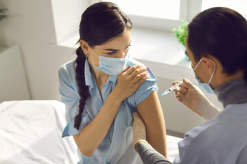 Young female patient in medical mask getting vaccination from 19-ncov infection from doctor or nurse in medical clinic. Vaccinating against COVID-19 infection concept
