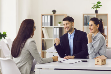 Joyful husband and wife with a realtor in the office sign a contract to buy a new home. Young family happily clenching their fists at buying a house or getting mortgage approval. Real estate concept.