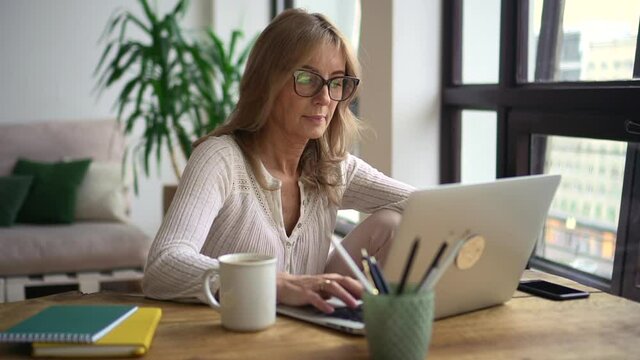 Mature woman sit at desk and work at laptop at home office. Entrepreneur agent look at info at screen. Confident 50s small business owner. Employee, project concept