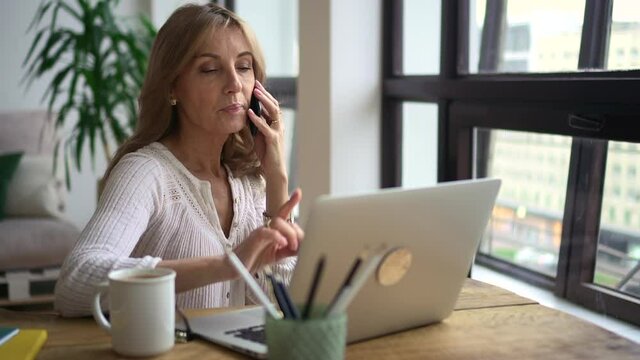 Mature business woman working on laptop and smartphone. Discuss business issues on phone. Female entrepreneur sit in office and talk with colleague, coworker. Concept computer
