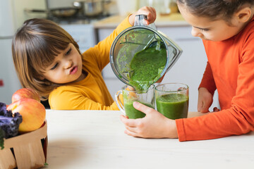 Happy children pour glass cup of green smoothie from blender. Cute boy and girl crazy drinks healthy dietary nutritious cocktail at home in the kitchen. Healthy lifestyle, raw food