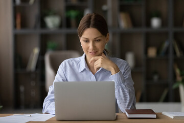 Smiling young Caucasian businesswoman sit at desk at home office look at laptop screen consult client online. Millennial female employee or woman worker work distant on computer, analyze paperwork.