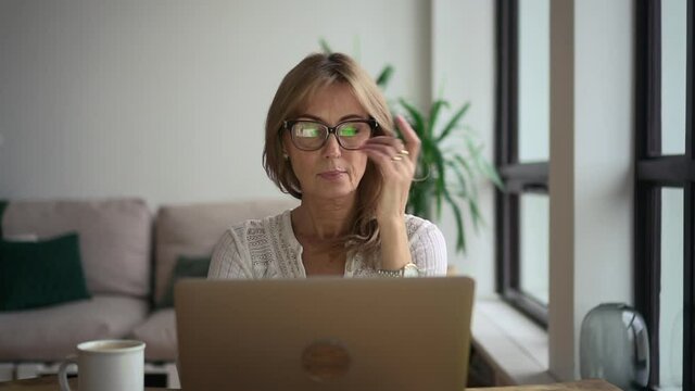 Businesswoman working with laptop and sitting at table in home office during pandemic. Front view of middle aged American woman looks at computer screen and types text, sits at desk in light room with