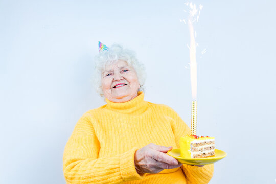 Older Woman With A Gift Wear Yellow Sweater And Horn Cap On A White Background Holding Plate With Cake With Fireworks