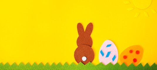 Easter holiday. Cut out of felt applications of two eggs and brown rabbit on the grass. Yellow background. Copy space. Flat lay. Banner