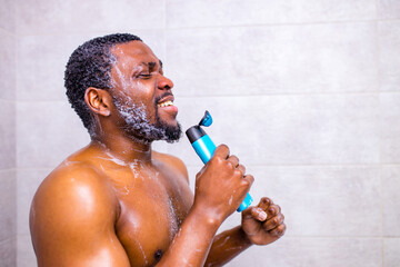 Fototapeta na wymiar Attractive young cheerful man singing while washing in the shower, holding shampoo bottle like microphone