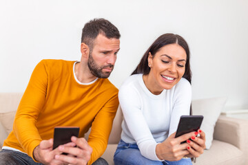 Young couple using smartphones at home. Jealous boyfriend spying his girlfriend's phone. Jealous...