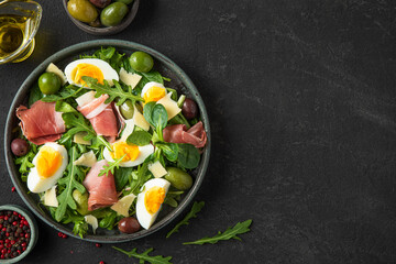 Healthy diet food. Salad with prosciutto, cheese, olives, eggs and arugula in a plate on dark background. Top view