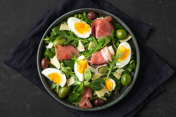 Prosciutto di Parma salad with olives, parmesan cheese, eggs and arugula on dark background. Top view