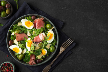 Prosciutto di Parma salad with olives, parmesan cheese, eggs and arugula on dark background....
