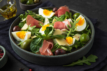 Salad with prosciutto, parmesan, olives, eggs and arugula in a plate on a dark background. Italian appetizer. close up
