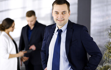 Fototapeta premium Portrait of a self-confident middle aged businessman in a blue suit, standing in a modern office with his colleagues at the background. Concept of business success