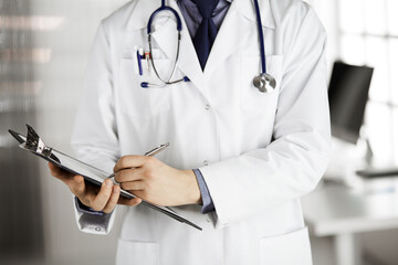 Unknown male doctor standing and working with clipboard of medication history records in clinic at his working place, closeup. Perfect medical service in hospital. Medicine and healthcare concept