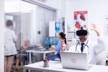 Fototapeta premium Portrait of professional caucasian male doctor in VR glasses sitting in hospital cabinet and making gestures using virtual reality innovations while female nurse working in background