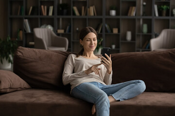 Obraz premium Millennial Caucasian woman sit relax on sofa in living room talk on video call on smartphone. Happy young female renter rest on couch at home use modern cellphone, have webcam virtual event online.