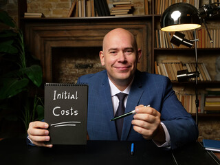 Man wearing blue business suit and showing black notepad with written text Initial Costs . Blurred...