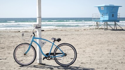 Fototapeta premium Blue bicycle, cruiser bike by ocean beach, pacific coast, Oceanside California USA. Summertime vacations, sea shore. Vintage cycle on sand near lifeguard tower or watchtower hut. Sky and water waves.