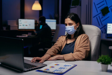 Stressed caucasian young entrepreneur doing overtime wearing face mask . Woman following social...