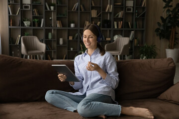 Obraz premium Smiling young Caucasian woman sit on sofa at home wear headphones talk on video call. Happy millennial female relax on couch have webcam digital conversation on pad. Virtual event concept.