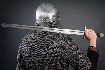 Back view of a knight in chain mail and a helmet with a sword on his shoulder on a dark background....