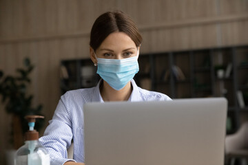 Fototapeta na wymiar Focused businesswoman in facemask look at laptop screen work online during coronavirus pandemics quarantine. Concentrated Caucasian female employee in facial mask use computer. Covid-19 concept.