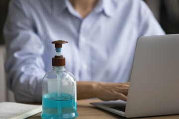 Close up of bottle with liquid sanitizer on desk in office, female employee work on computer in background. Antibacterial gel protect from coronavirus covid-19 pandemics at workplace. Corona concept.