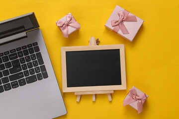 Laptop, empty chalk board and gift boxes on yellow background