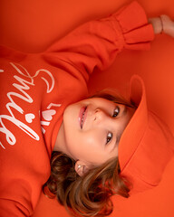 Smiling little child girl 6-7 years old in cap for mock up and fashionable clothes lies on floor in studio on orange background. Copy space
