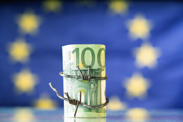 European Union currency wrapped in barbed wire against flag of EU as symbol of Economic warfare, sanctions and embargo busting.