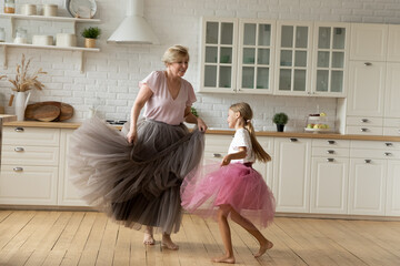 Fototapeta Merry leisure time. Happy energetic grandmother teach ball dance active little child. Caring grandma junior girl grandkid engaged in dancing in funny large fluffy skirts barefoot on warm kitchen floor obraz