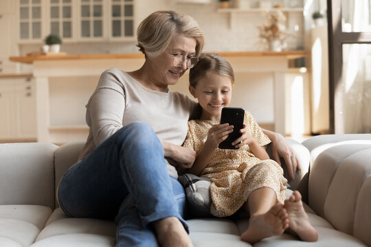 Mutual interests. Happy small girl and older grandmother embrace on cozy couch at living room look at cell screen surf web shopping online. Aged grandma spend time with beloved grandchild using gadget - Powered by Adobe