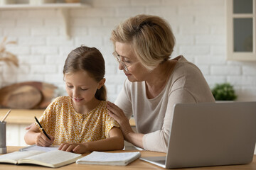 Loving senior grandmother hug shoulders of small girl grandchild assist in preparing homework support give advice. Friendly aged woman teacher watching little kid pupil writing maths problem solution